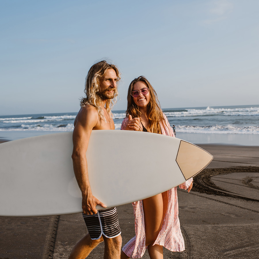 happy-couple-walking-together-on-beach-with-surfin-SUWC9HH.jpg